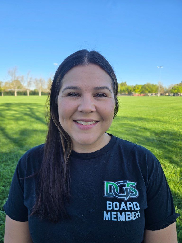 Man in green polo stands on a football field, arms crossed, stadium in background.