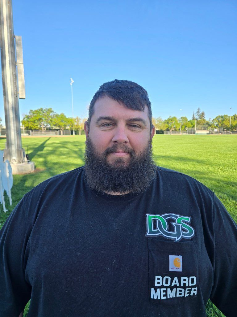 Football coach with arms crossed, looking stern on a sunny field, players in the background.