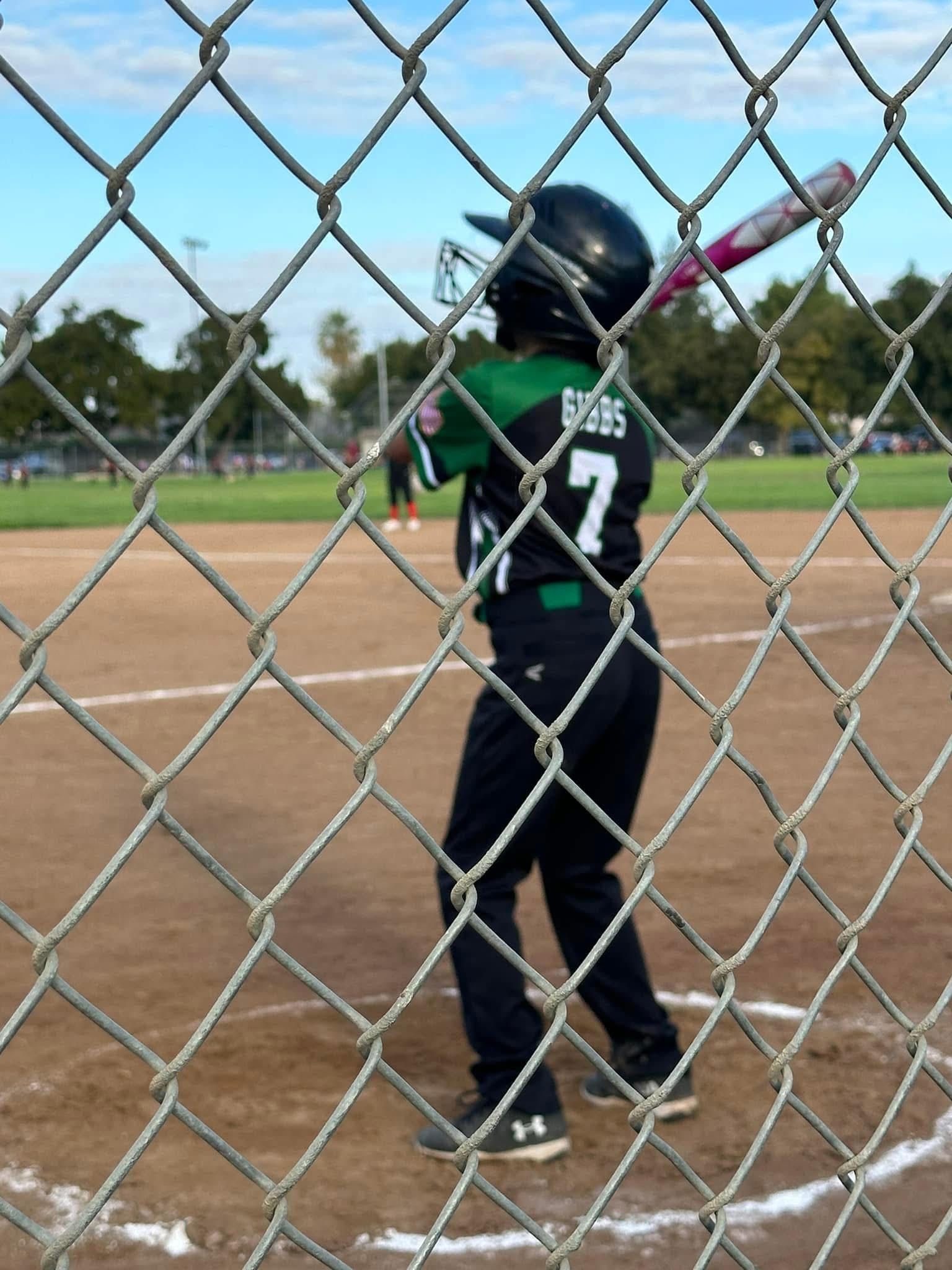 A person wearing a hat and orange shirt, holding a baseball bat on their shoulder on a baseball field.
