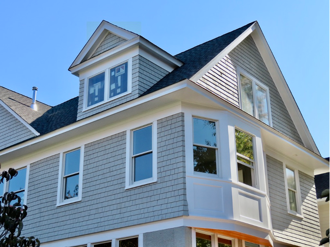 Gray house with multiple windows, bay window, and a gabled roof against a blue sky.