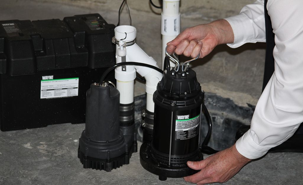 Man holds a black sump pump, next to another and battery box in a basement setting.