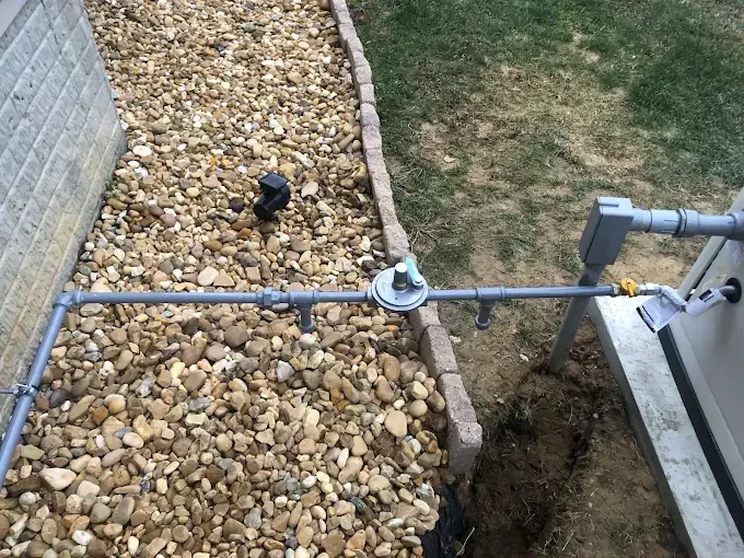 Pipes and a valve on a gravel bed near a wall and lawn, with part of a machine on the right.