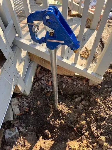 Blue frost-free hydrant installed near a white deck, surrounded by dirt and leaves.