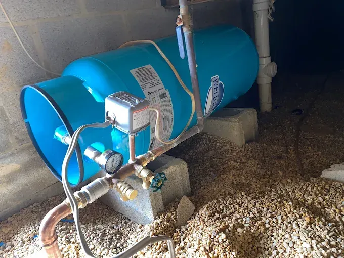 Blue water pressure tank, copper pipes, and a pressure gauge set up in a crawlspace on concrete blocks.