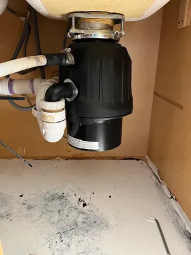Under-sink view: A black garbage disposal is connected to white pipes and the sink drain.