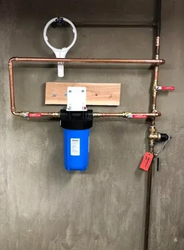Water filter system with copper pipes, blue filter, shut-off valves, on a concrete wall.