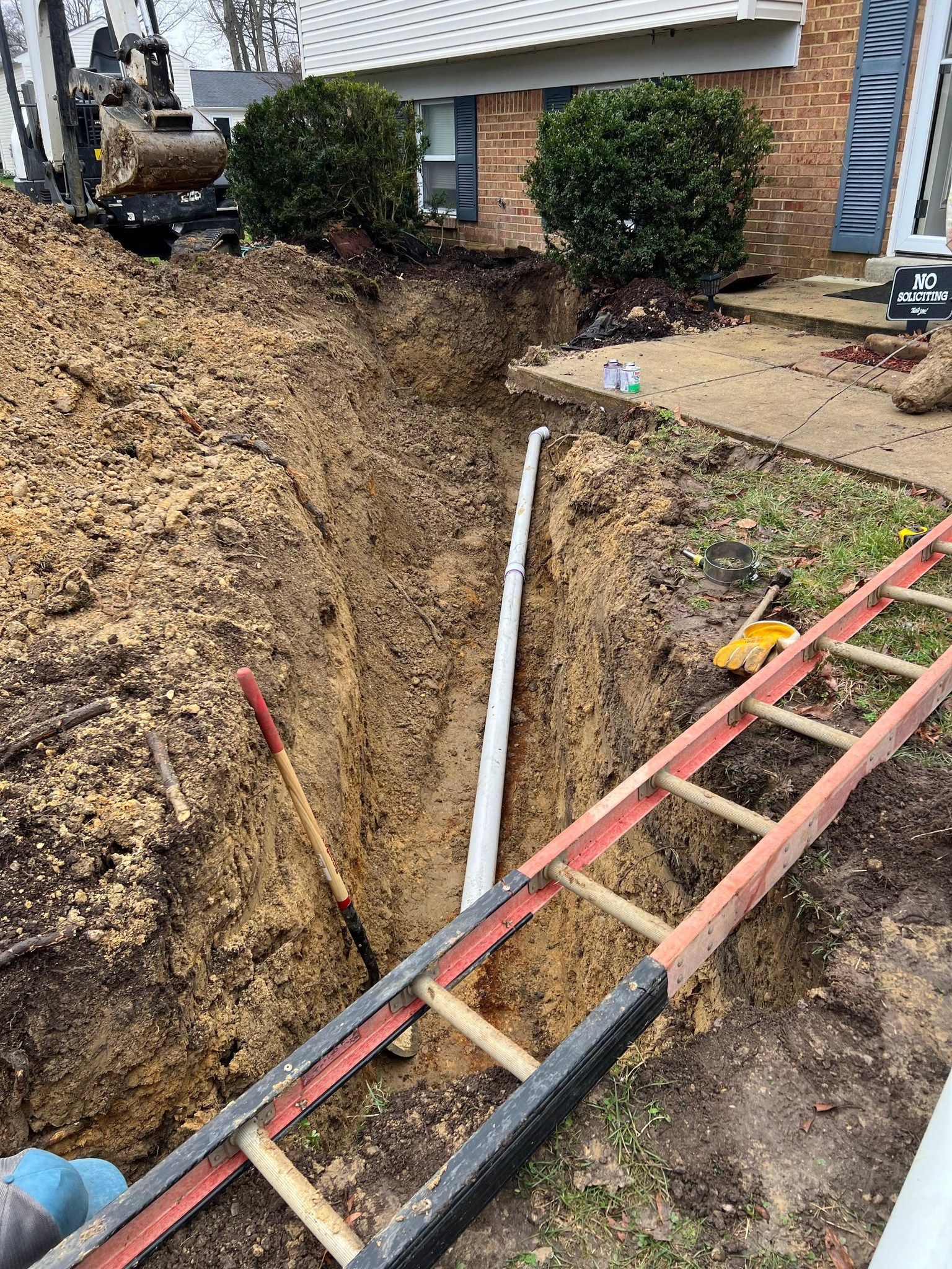 Trench dug in front yard with white pipe, ladder, and tools; near a brick house with bushes.