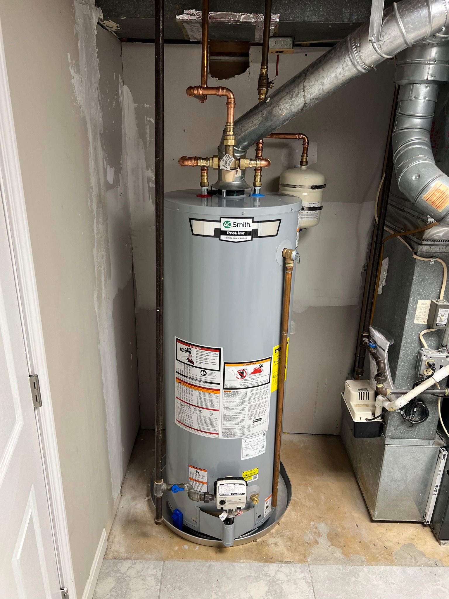 A water heater is installed between a wall and a furnace. Metal pipes and tubes are connected to the top.