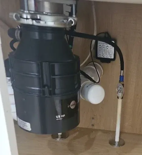 Black garbage disposal under a kitchen sink, connected to plumbing and electrical components.