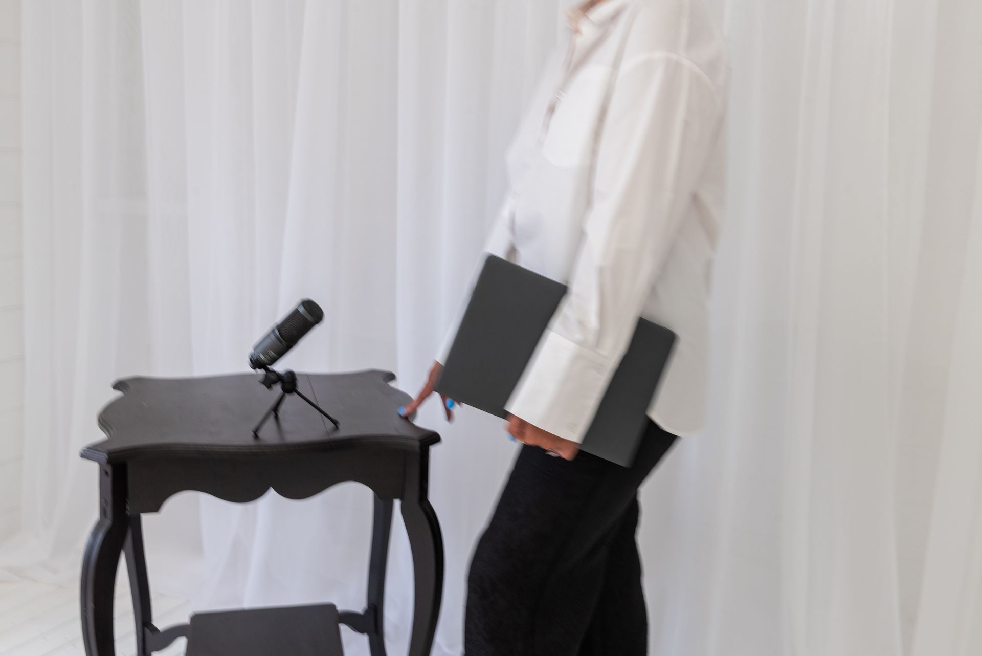 Person in white shirt and black pants holding a dark notebook, standing near a table with a microphone. White backdrop.