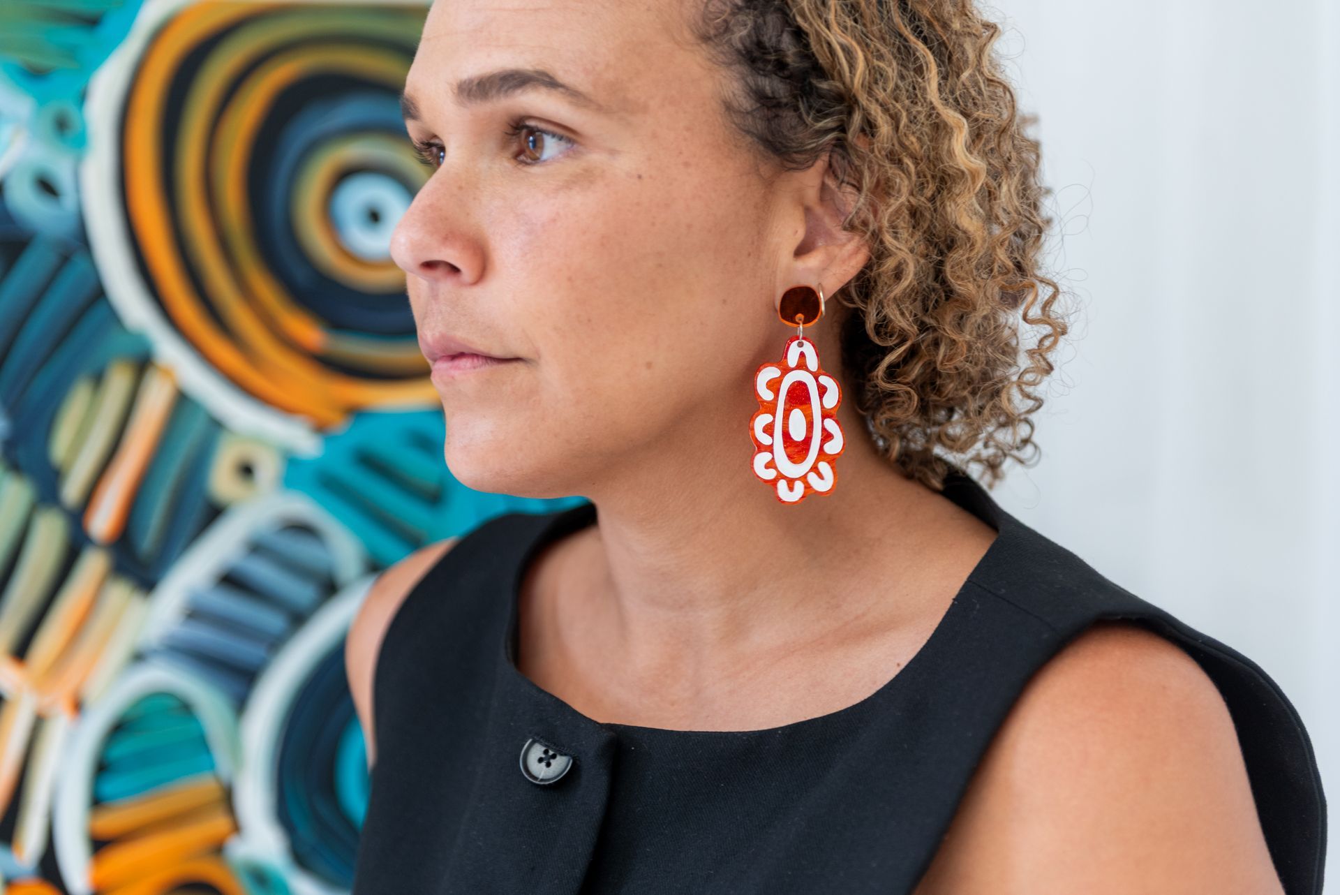 Woman with curly hair wearing red and white earrings, looking to the side near a colorful painting.