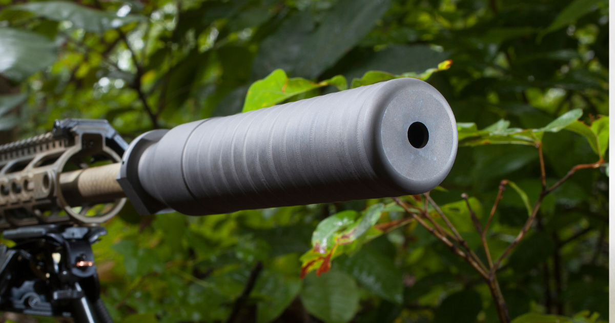 A grey rifle suppressor attached to a rifle in a wooded environment.