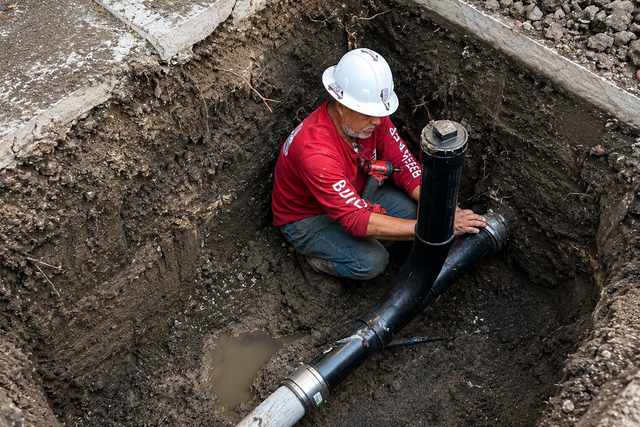 Worker in a hard hat repairs a black pipe in a muddy trench.