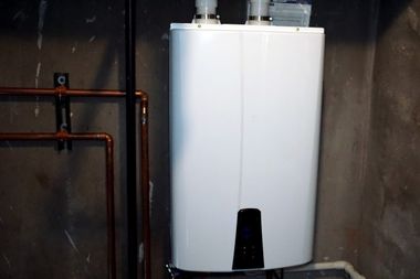 White wall-mounted water heater in a basement with copper pipes on the left and dark concrete walls.