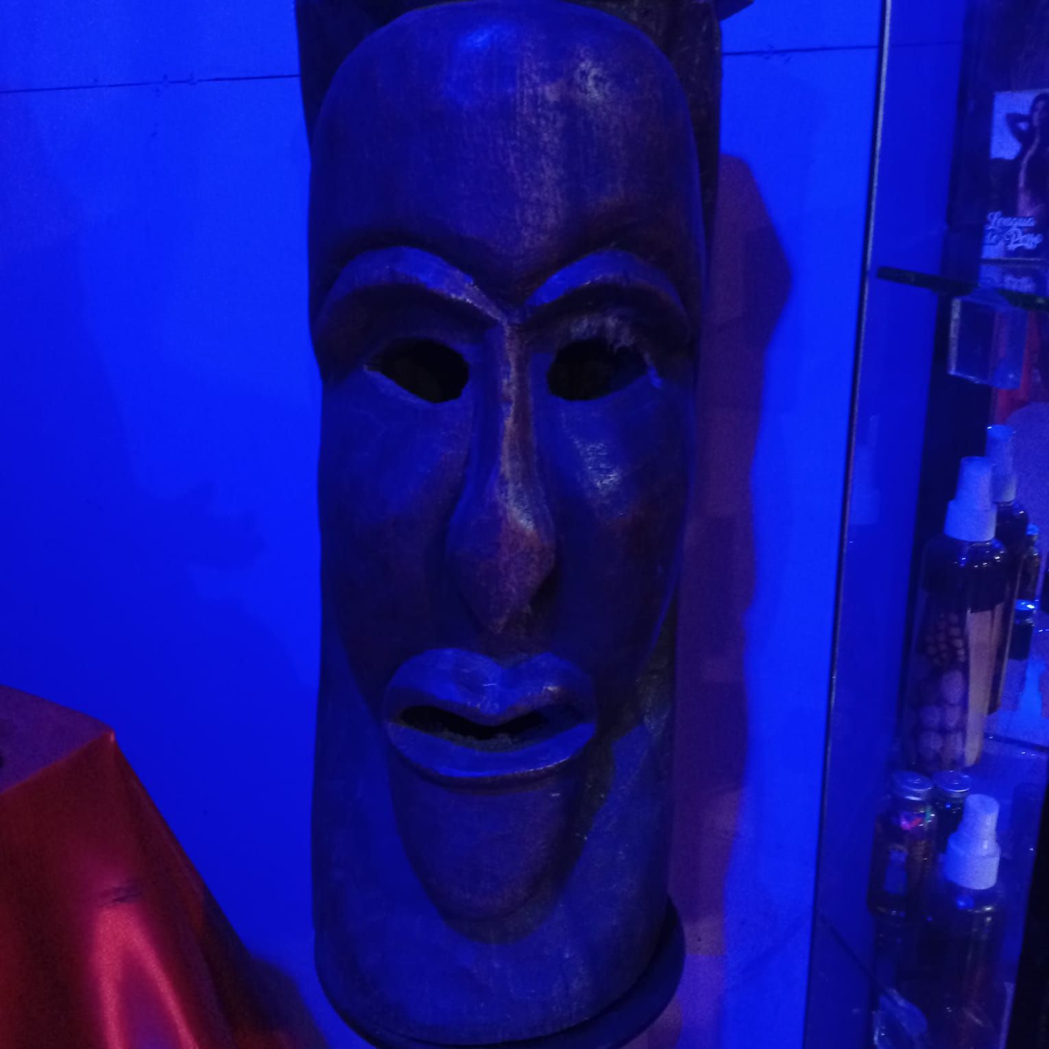 Máscara africana de madera, enmarcada centralmente, contra una pared iluminada con luz azul. La máscara es de color marrón oscuro.