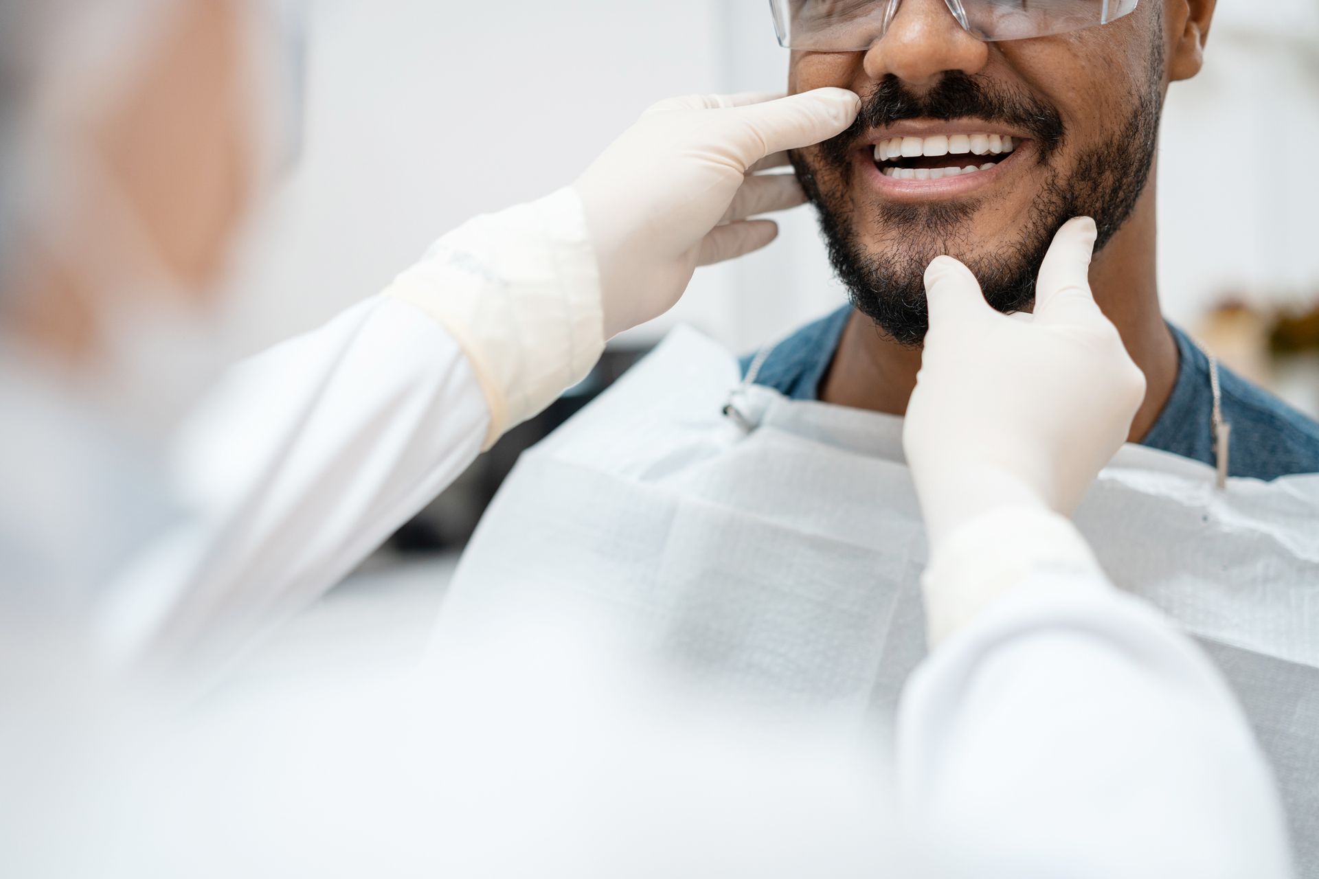Dentist examining a Black man's teeth in a dental chair. Man smiles as dentist holds his cheeks.