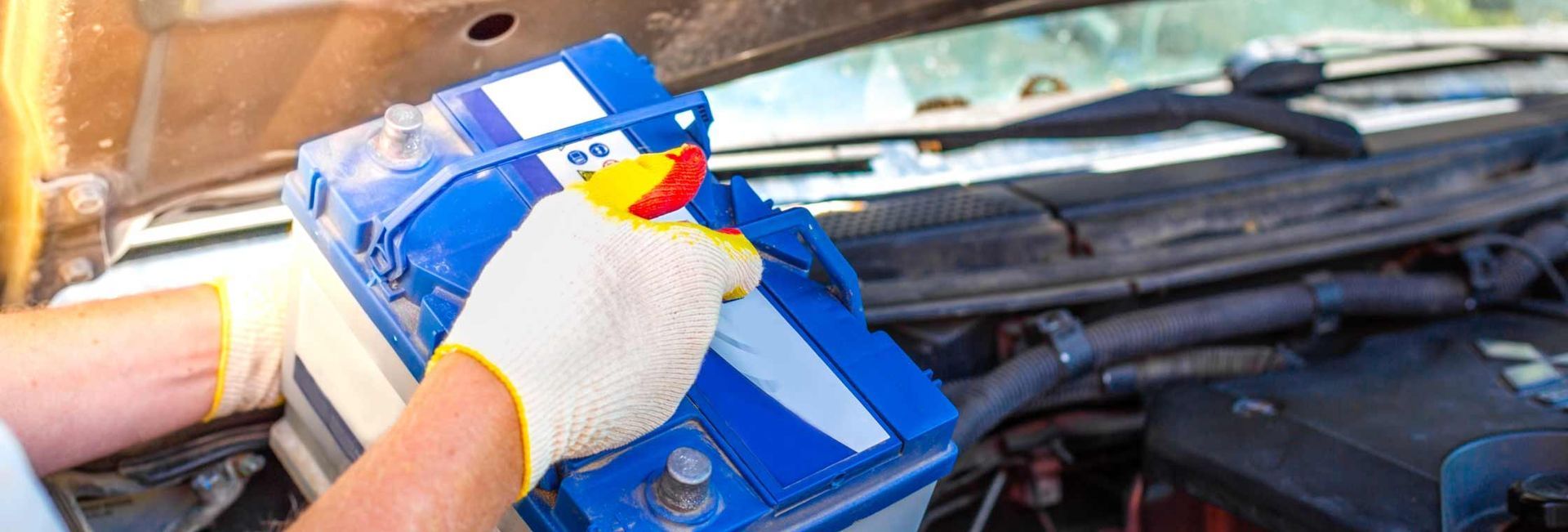 A person wearing gloves is holding a blue car battery under the open hood of a car.