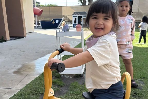 Smiling girl riding a yellow tricycle at the Montessori outdoor play area.