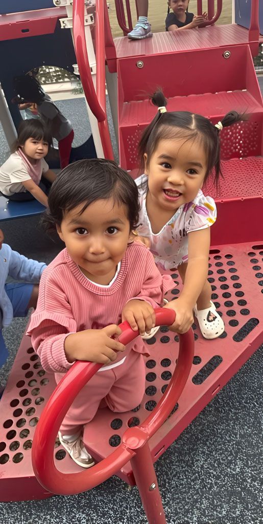 Two toddlers smiling on red playground equipment at Montessori school