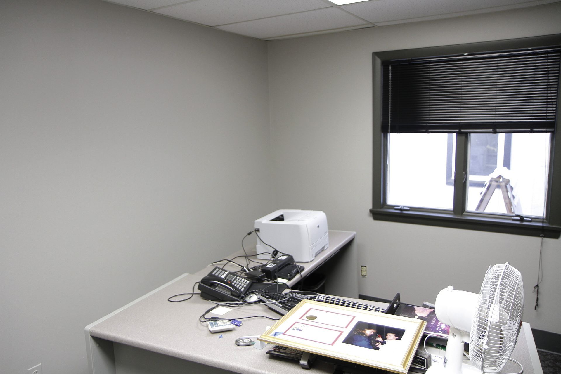 A desk with a printer and a fan in a room with a window.