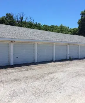 A row of white garage doors with a gray roof in a parking lot.