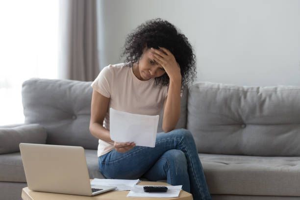 Person sitting on couch reviewing bills with laptop and documents on coffee table.