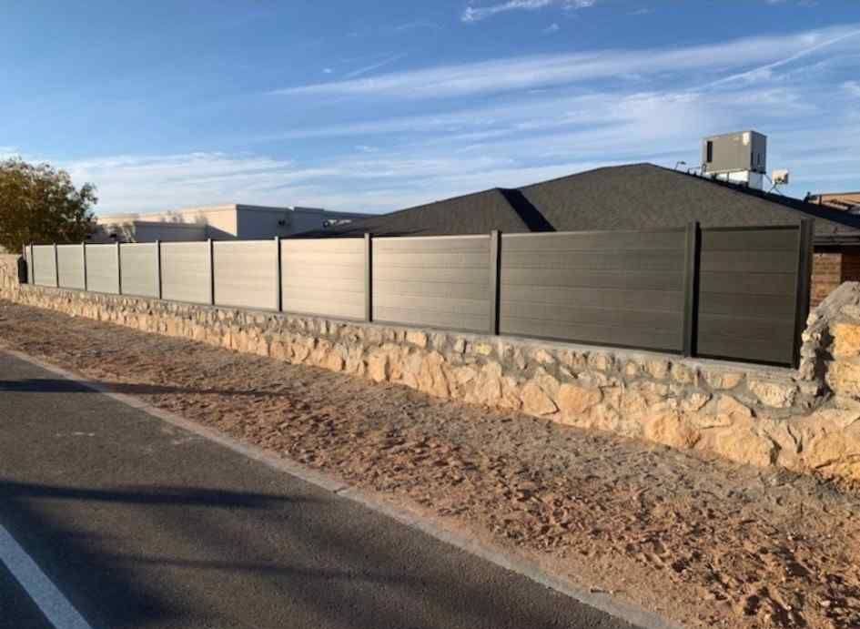 Desert roadside with a stone wall and dark fence in front of a modern house under a blue sky