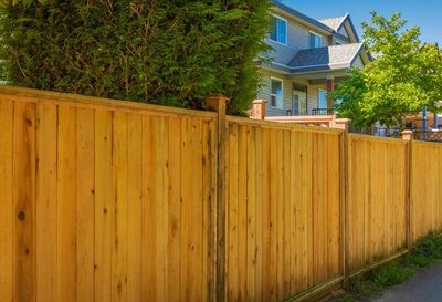 Wooden fence in front of a house, partially obscured by trees, under a blue sky.