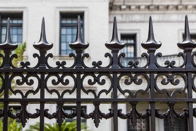 Black wrought iron fence in front of a white building with dark windows and a balcony.