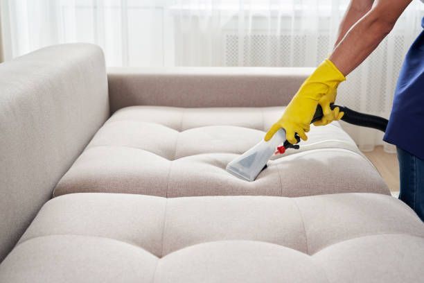 Person in yellow gloves cleaning a beige tufted couch with a vacuum.