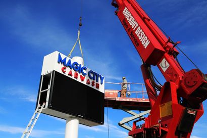 Crane lifts a Magic City sign onto a pole against a blue sky.