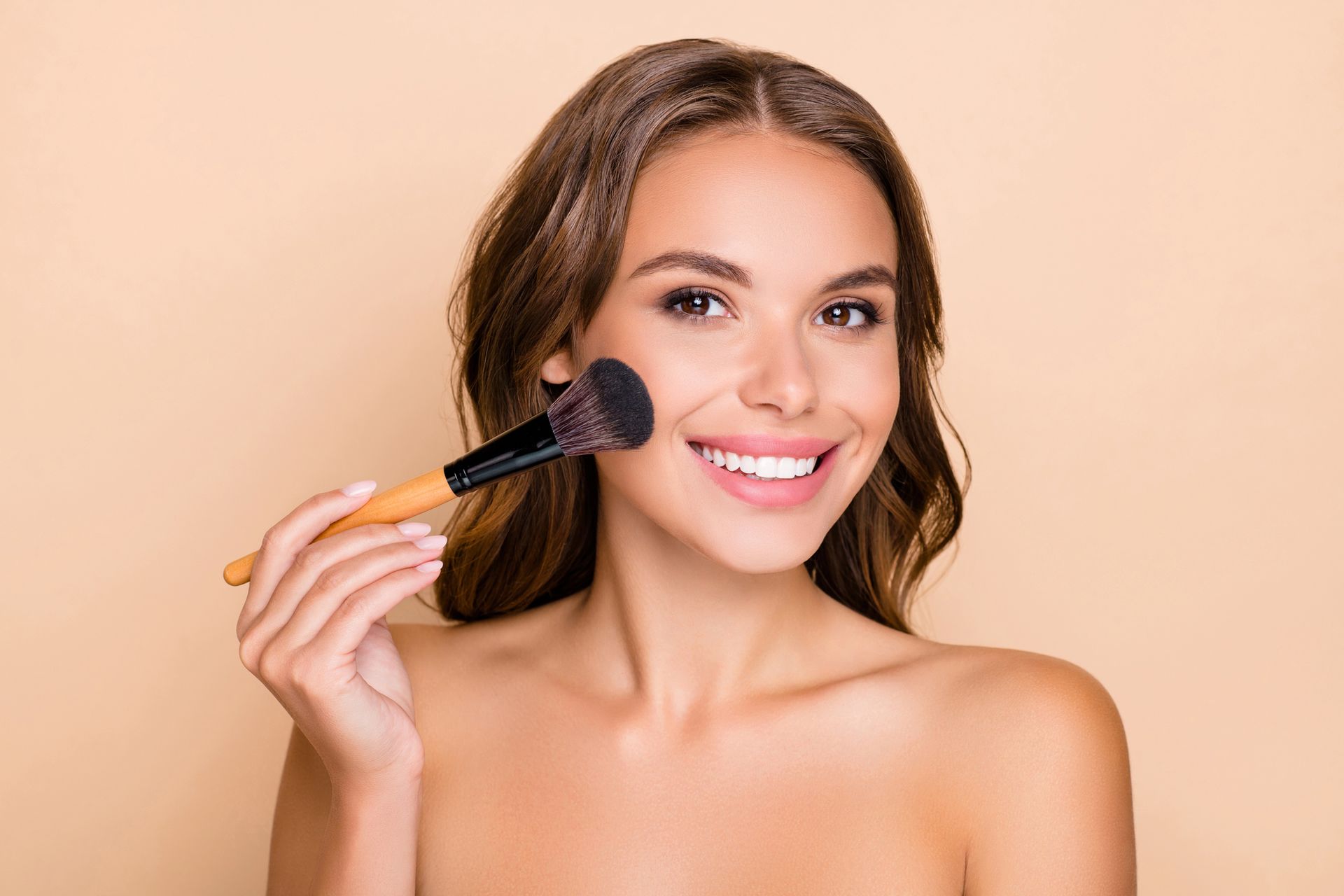 Mujer sonriendo, aplicando rubor con una brocha de maquillaje, fondo bronceado.