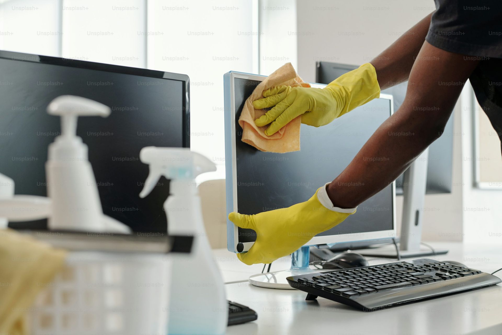Person in yellow gloves cleaning a computer monitor with a cloth; office setting.