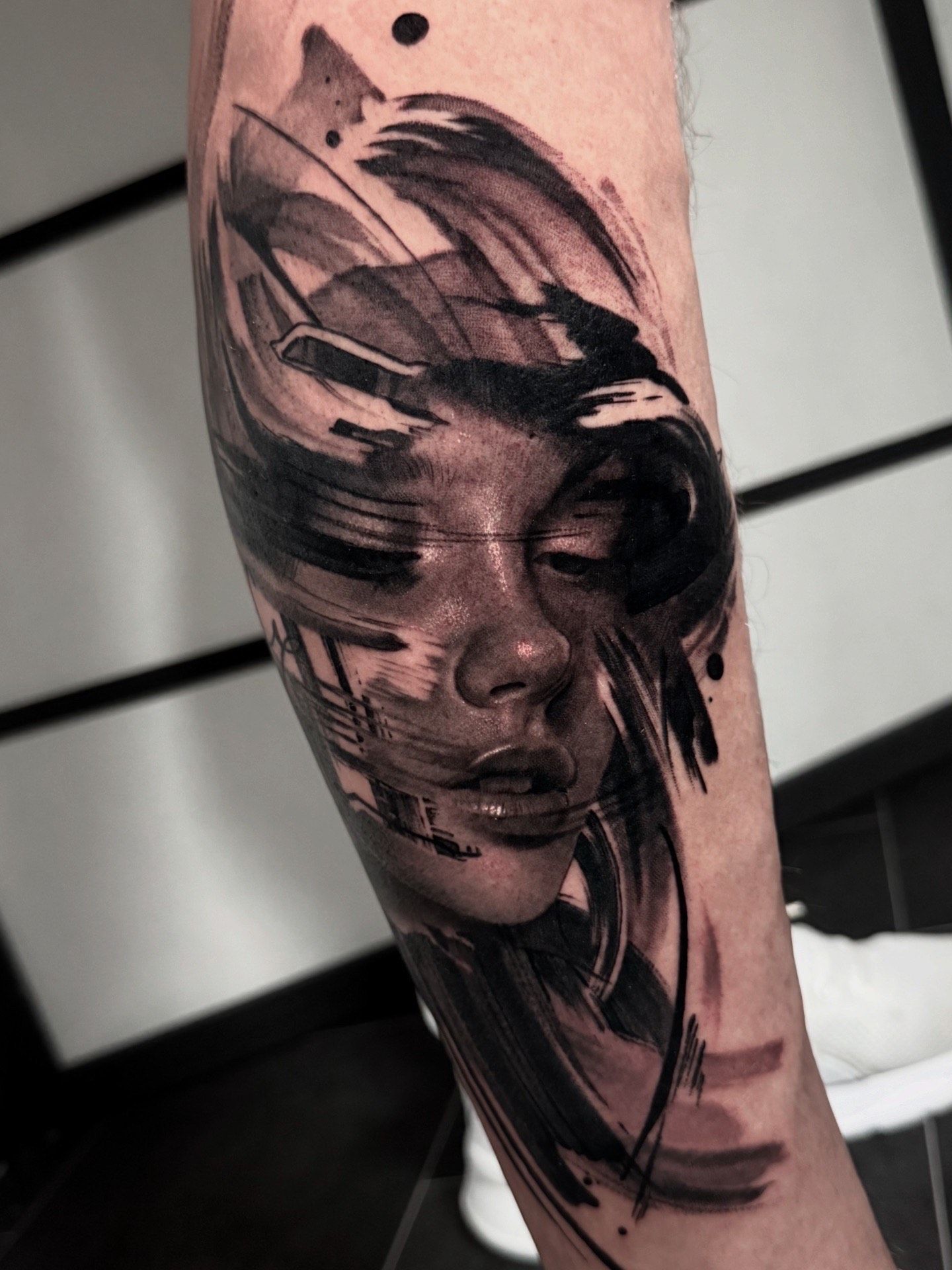 A close-up of an arm tattoo featuring a hyper-realistic face dissolving into dark, abstract brushstrokes and black ink.
