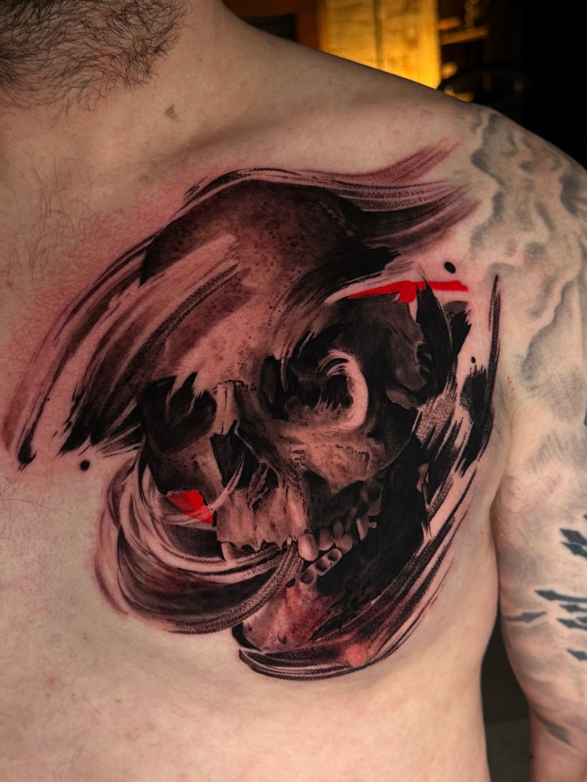 A chest tattoo of a skull rendered in a painterly, abstract style with black, grey, and bold red brushstroke accents.
