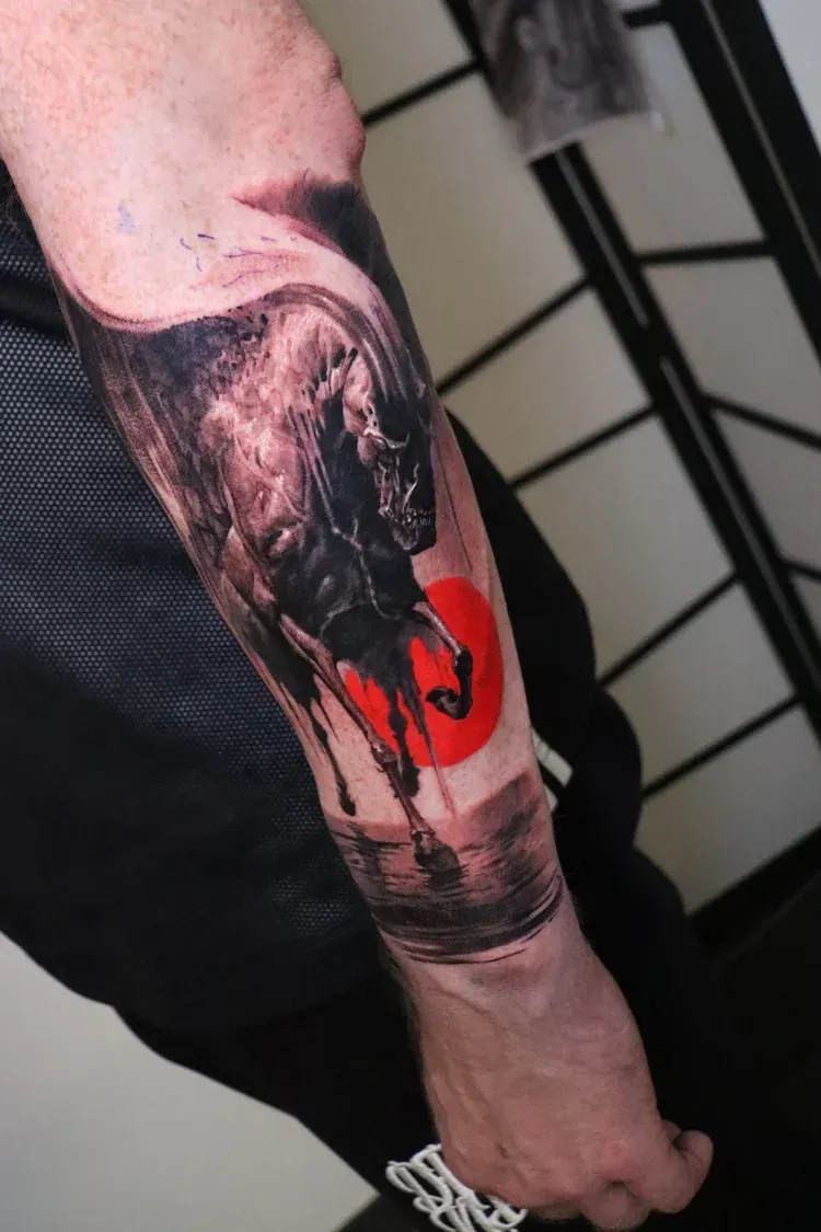 A forearm tattoo featuring a dark, textured bull silhouette standing before a bold, circular red sun.