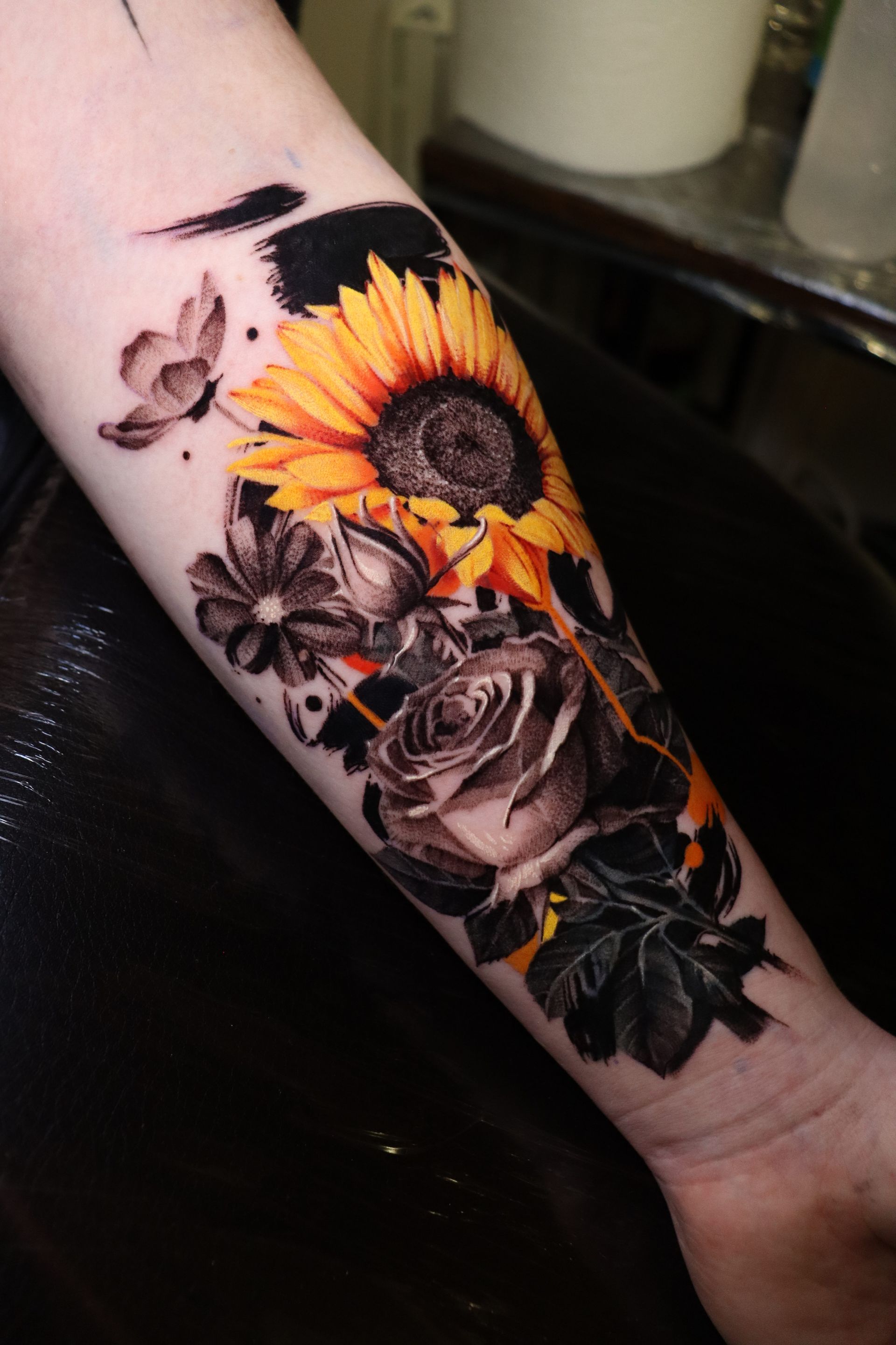 Forearm tattoo featuring a bright yellow sunflower contrasted with a black-and-grey rose and abstract black ink strokes.
