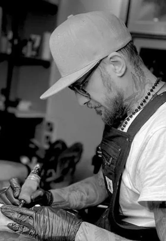 A tattoo artist in a cap, glasses, and apron uses a tattoo machine to work on a client’s arm in a studio.