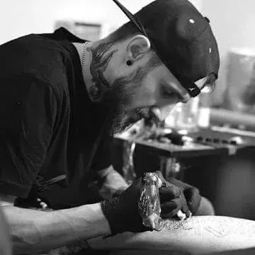 A person with neck tattoos and a backward cap focused on tattooing a person's arm while wearing black gloves.