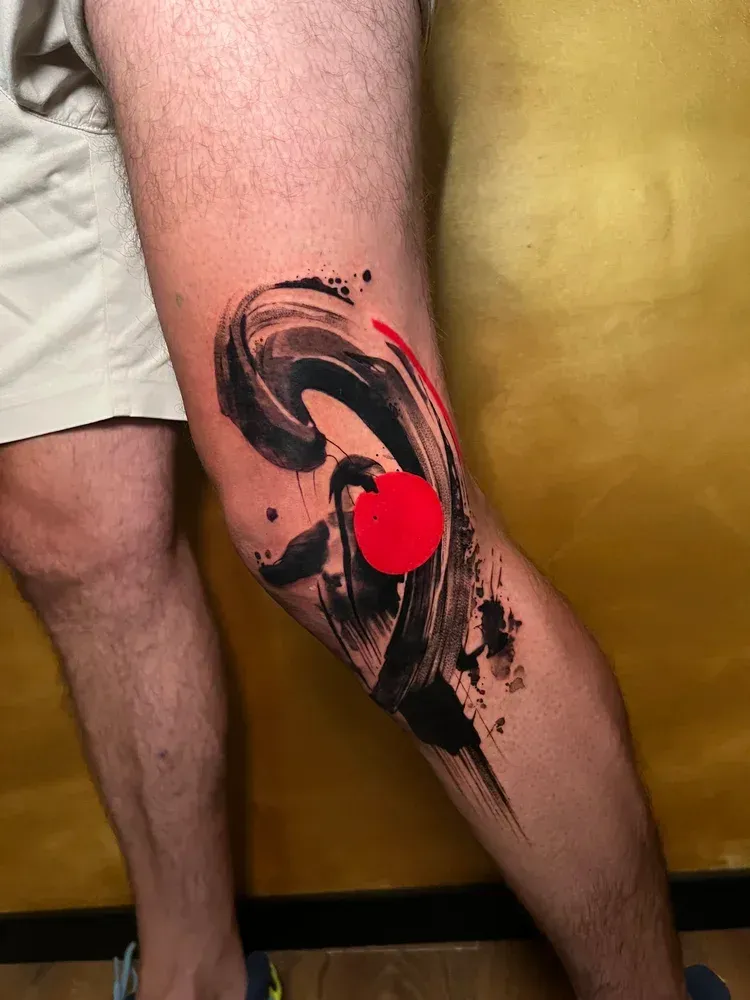 A knee tattoo featuring a black, brushstroke-style circular design accented by a bright red sun-like solid circle.