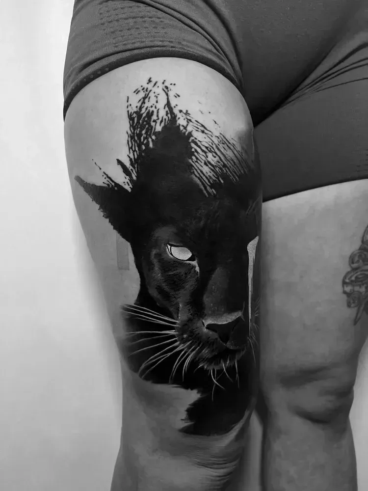 A black and white tattoo of a panther head on a person's upper thigh, featuring abstract brushstroke effects.