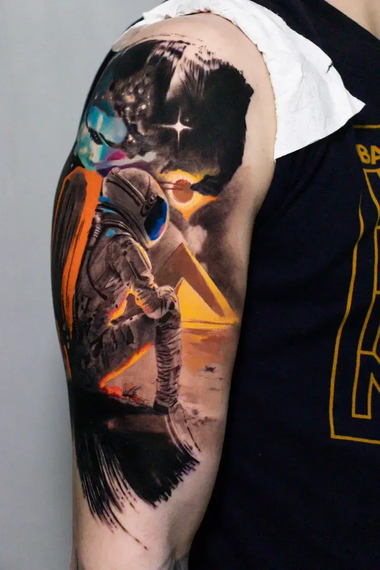 A colorful tattoo on an upper arm depicting an astronaut sitting on sand before a pyramid under a starry space sky.