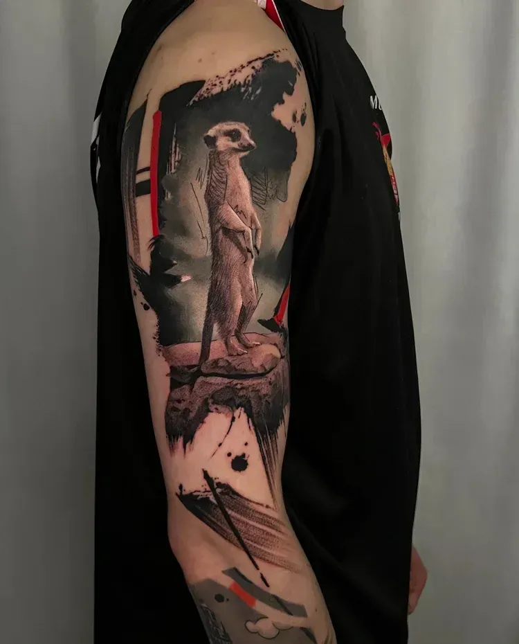 A tattoo of a meerkat standing on a rock, rendered in a black and grey style with abstract ink-wash accents on an arm.