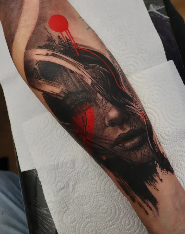 A forearm tattoo featuring a portrait with sweeping black brushstrokes, vibrant red accents, and a red circular emblem.