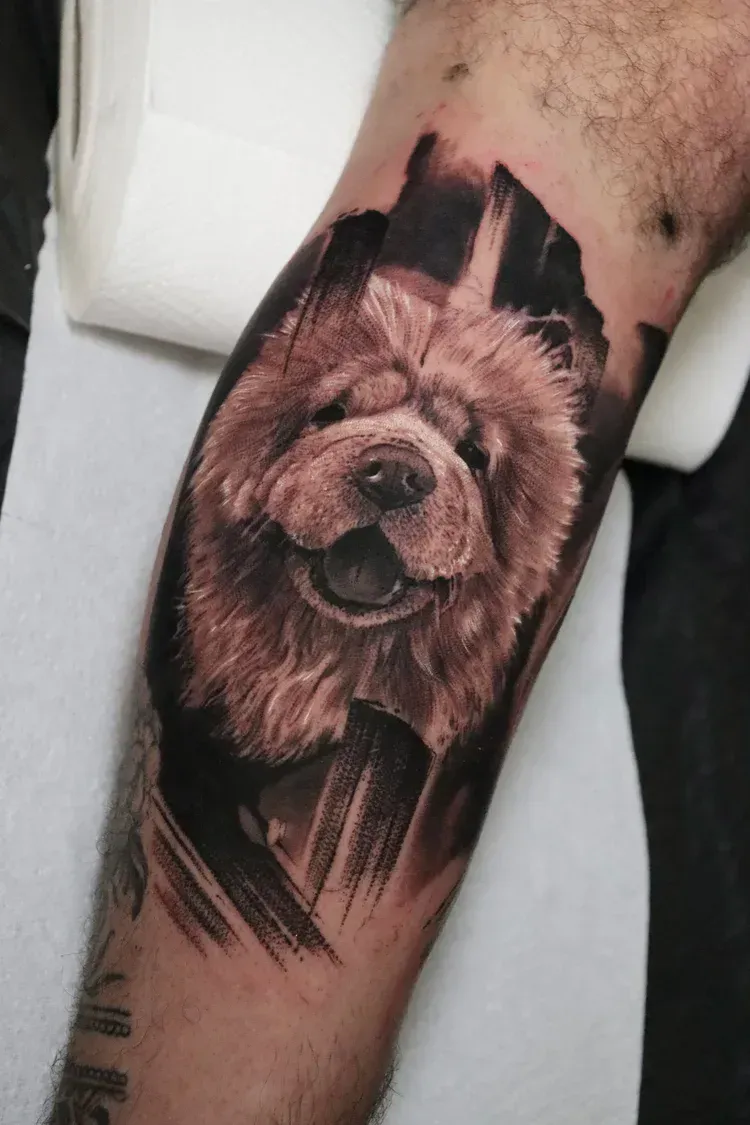 A realistic portrait tattoo of a fluffy Chow Chow dog on a person's lower leg, surrounded by dark, abstract brushstrokes.