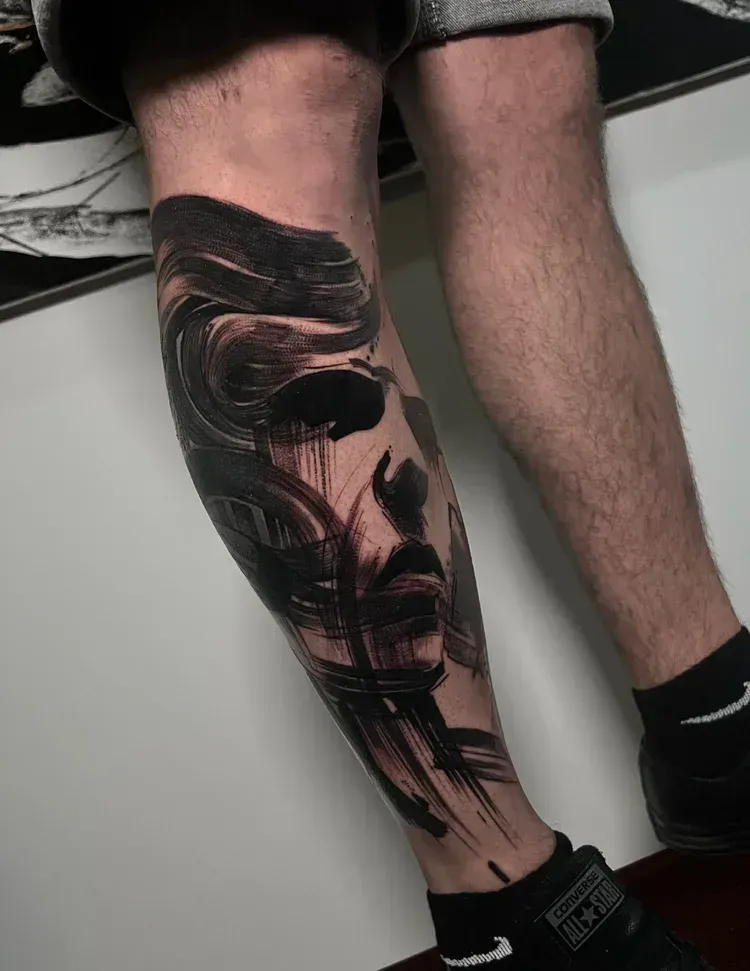 A lower leg features an abstract black ink tattoo resembling brushstrokes forming a human face in a dark, artistic style.