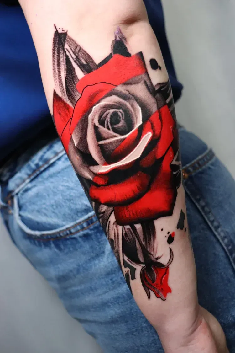A tattoo on a forearm featuring a detailed rose with deep red petals and black-and-grey shading and ink splatter accents.