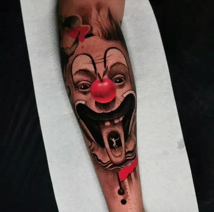 A detailed tattoo on an arm depicting a clown with a 3D red nose, a wide mouth, and a tiny figure visible inside its throat.