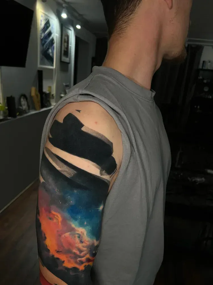 A person wearing a grey sleeveless shirt, showcasing a sleeve tattoo with bold black brushstrokes over a colorful nebula.