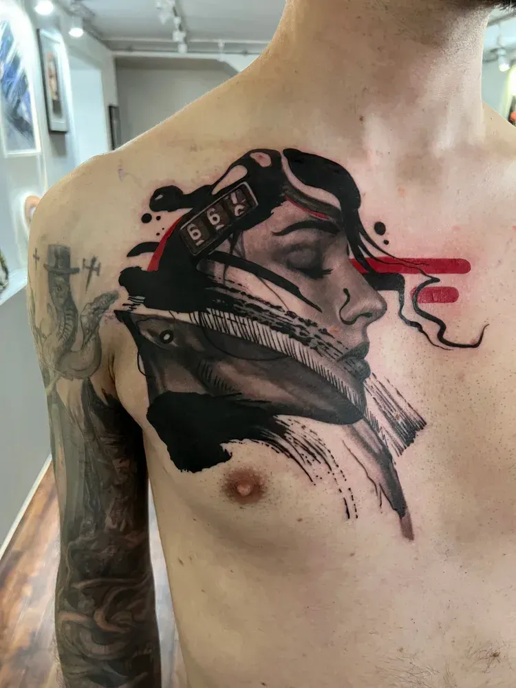 A tattoo on a chest featuring a profile face with abstract black ink splashes and horizontal red lines.